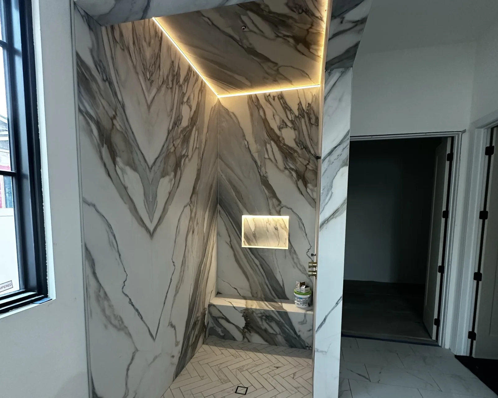 Bookmatched Calacatta marble walk-in shower with LED cove lighting, lit niche, marble bench, and herringbone floor by KV Tileworks